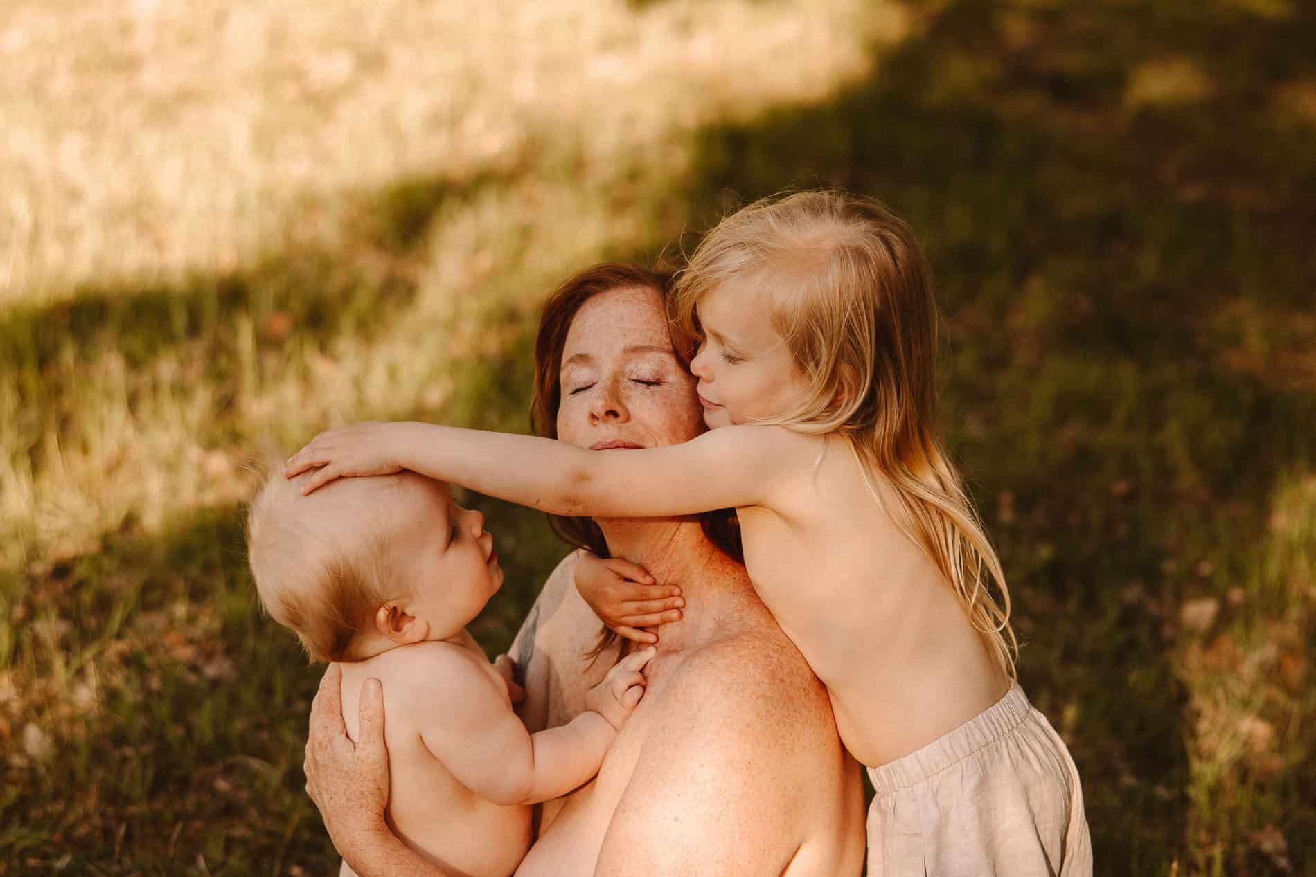 Familiebilde med mor og to barn som klemmer i utendørs omgivelser.