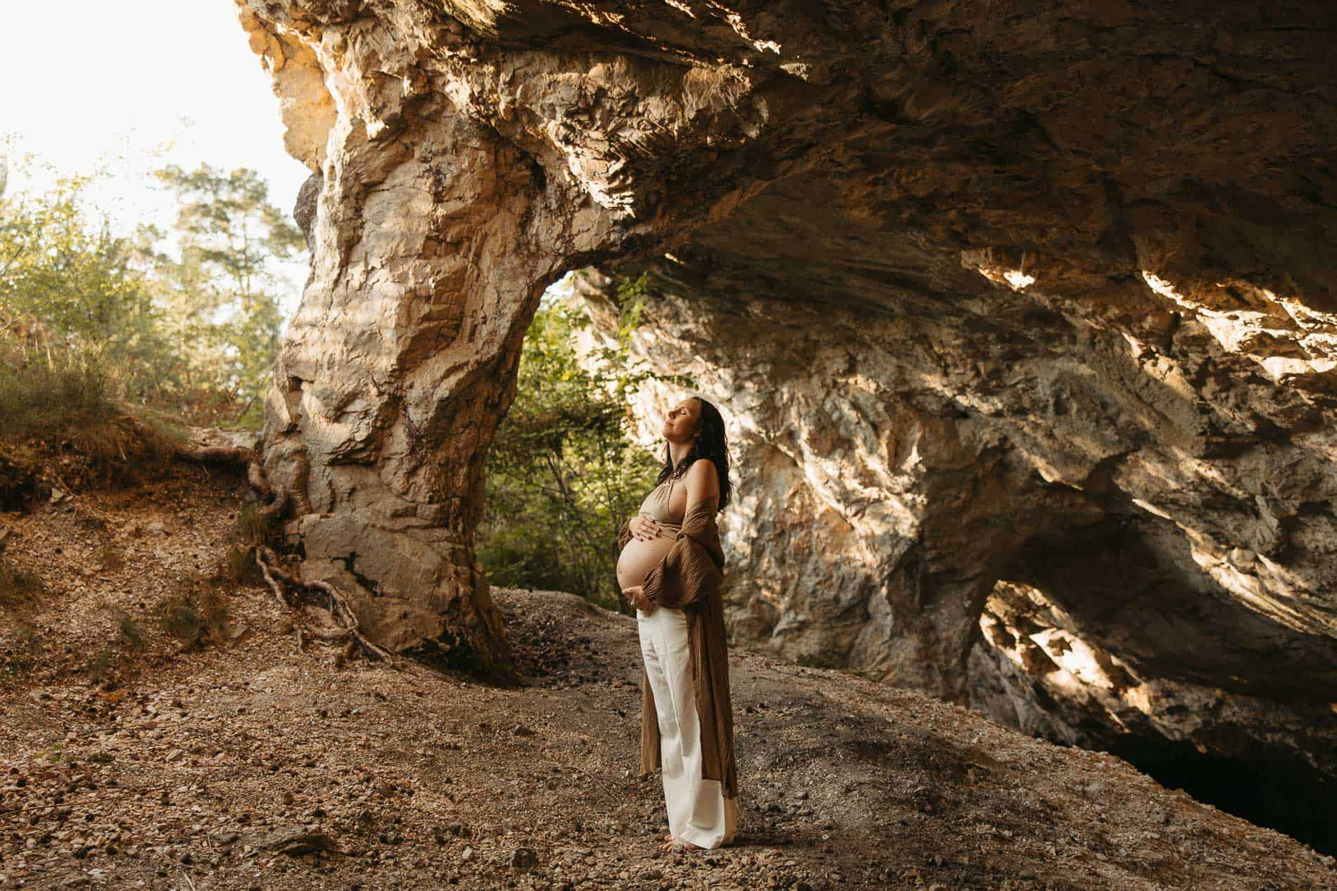 Gravide kvinne står under en stor steinåpning i naturen, omgitt av trær og stein.