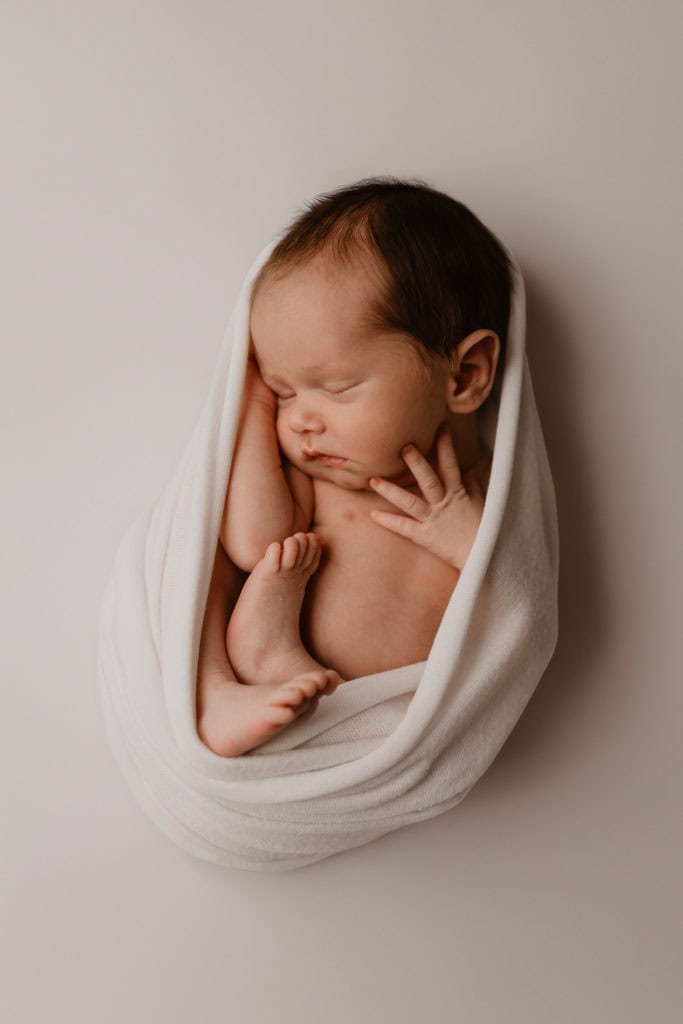 Nyfødt baby sovende, innpakket i hvitt teppe, på en nøytral bakgrunn.