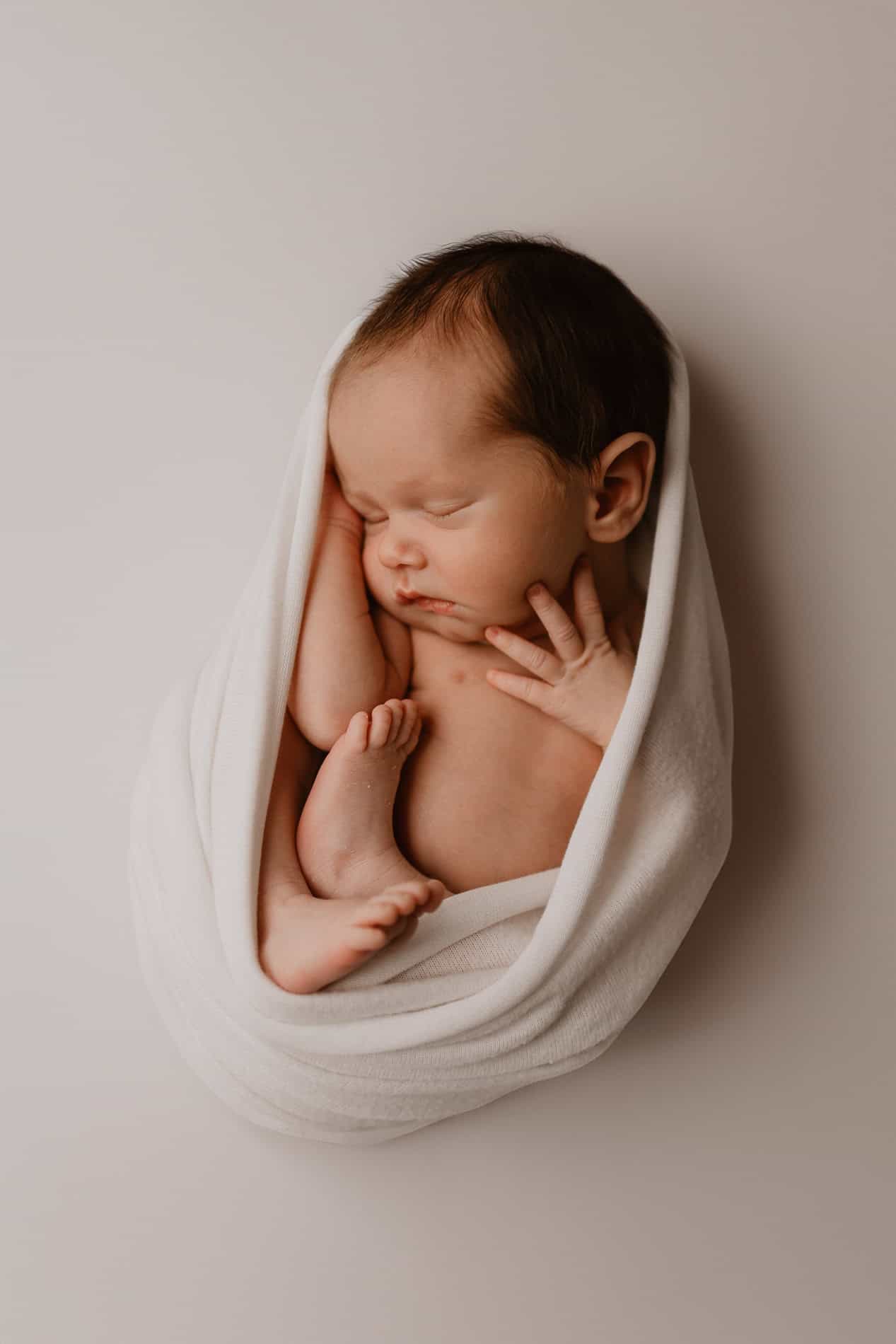 Nyfødt baby sovende, innpakket i hvitt teppe, på en nøytral bakgrunn.