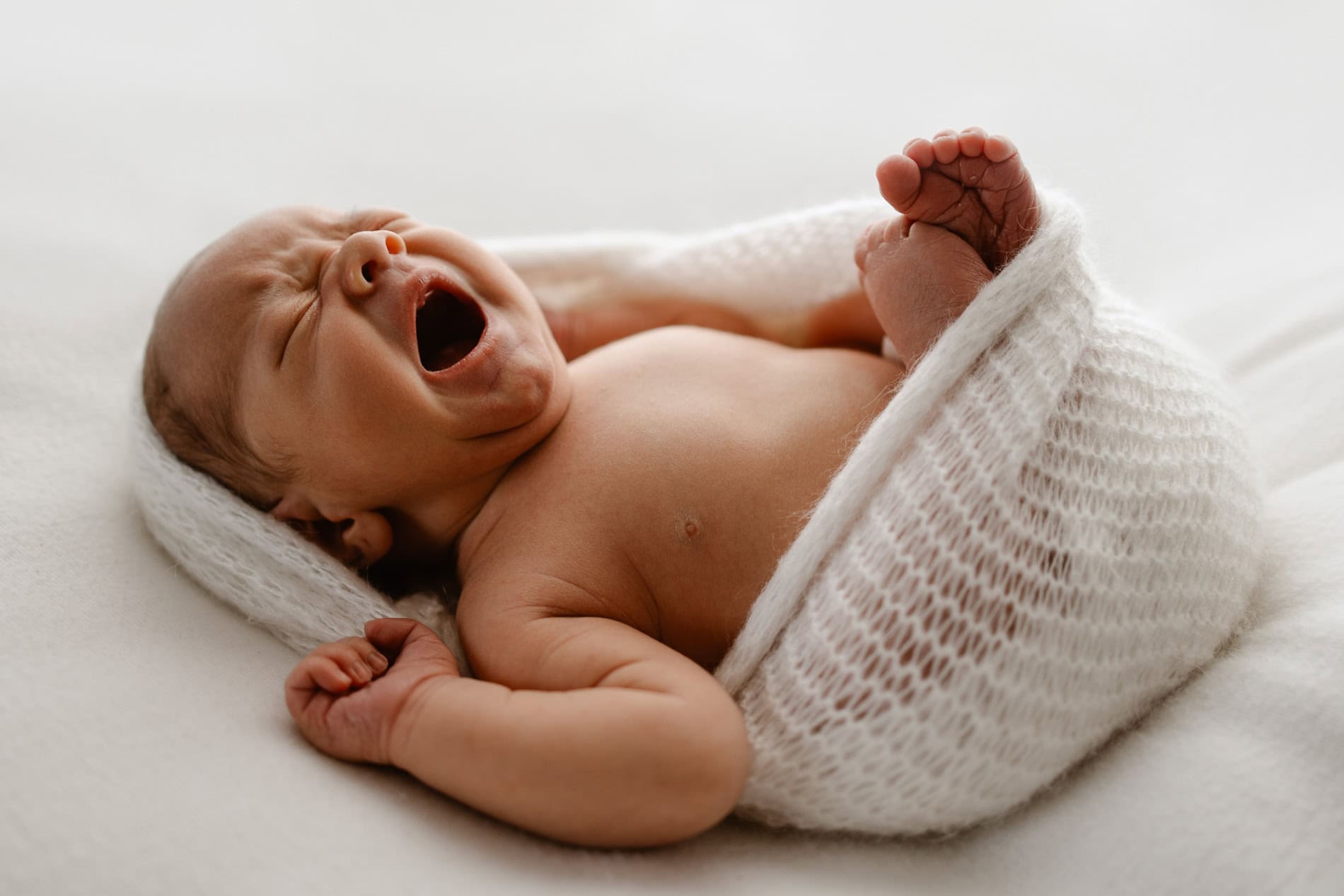 Nyfødt baby som sover, pakket inn i hvit strikket teppe på seng.